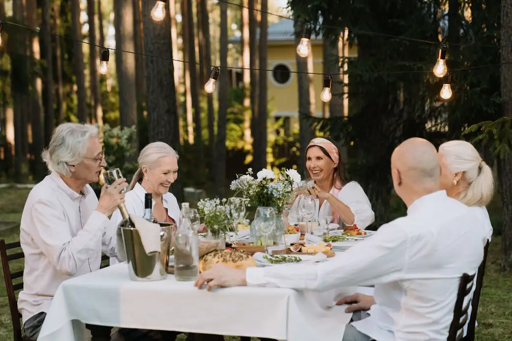 Five adults laughing and talking around a garden table with wine glasses and flowers under string lights