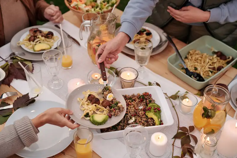 Hands serving food at a candlelit outdoor dinner table with pasta, salad and a pitcher of lemonade