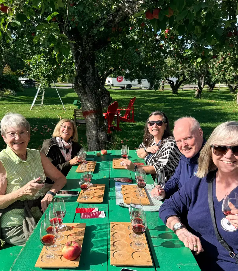 Five members tasting flights of wine at a green picnic table under an apple tree on the 2026 Quebec City trip