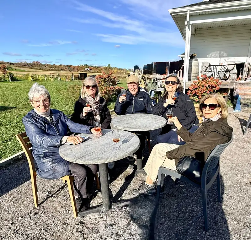 Five members raising glasses at patio tables outside a Quebec winery on a sunny autumn day